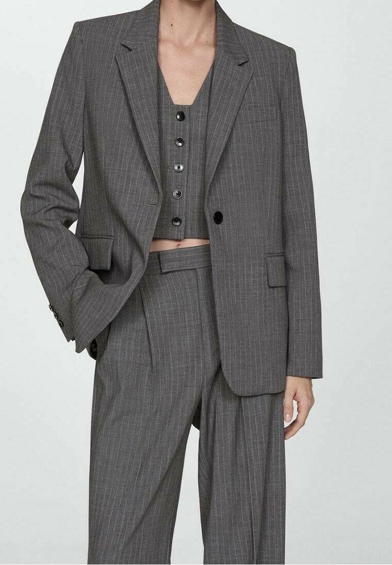 Frau trägt einen grauen Nadelstreifen-Dreiteiler mit hoch taillierter, weit geschnittener Hose, geknöpfter Weste und übergroßem Blazer vor einfachem Hintergrund.