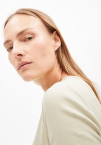 Top verde chiaro a costine con scollatura rotonda. Il soggetto ha capelli lunghi e lisci e guarda leggermente di lato. Sfondo minimo.