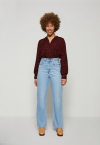 Camisa de botão de padrão floral vermelho com mangas longas, emparelhada com calças de ganga de perna larga em azul claro de cintura alta e sapatos de plataforma castanhos.