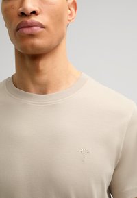 Beige katoenen t-shirt met een ronde hals, voorzien van een subtiele geborduurde bloemenontwerp op de linkerkant van de borst. Soepele textuur met korte mouwen.