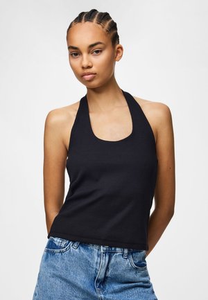 Jeune femme aux cheveux tressés portant un haut dos nu noir et un jean bleu, debout devant un fond blanc uni.