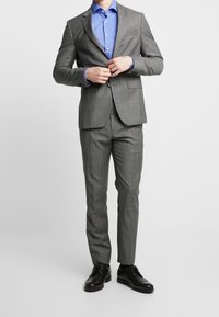 Šedý oblek na míru s dobře padnoucí bundou a úzkými kalhotami. Nošeno přes světle modrou košili. Černé společenské boty dokreslují celkový vzhled.