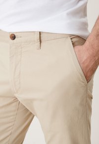 Homme portant un pantalon beige et une chemise blanche, avec la main droite dans la poche avant, montrant les détails du bouton et des passants de ceinture sur le pantalon.