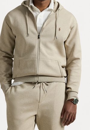 Hombre con sudadera beige con cremallera y pantalones deportivos a juego, con una camiseta polo blanca debajo y un reloj de pulsera marrón en la muñeca izquierda.