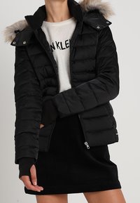 Czarna kurtka puchowa z kapturem wykończonym sztucznym futrem, zapinana na zamek, z ribbanym wykończeniem rękawów; noszona na lekkim swetrze i czarnej spódnicy.
