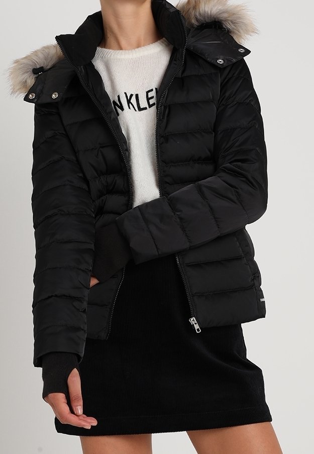 Czarna kurtka puchowa z kapturem wykończonym sztucznym futrem, zapinana na zamek, z ribbanym wykończeniem rękawów; noszona na lekkim swetrze i czarnej spódnicy.