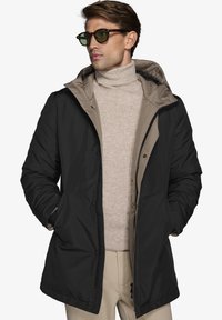 Cappotto isolante nero e beige con zip frontale, due tasche laterali e cappuccio, indossato sopra un maglione beige a collo alto. Occhiali da sole verdi.