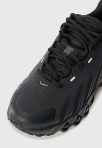Svart atletisk sneakers med texturerad mesh-övre del, böjd gummitåskydd, tjock snörning och dämpad sula med cirkulära detaljer.