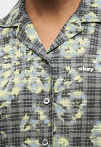 Detailný záber na károvanú košeľu so zeleno-modrým kvetinovým vzorom, zapínanú na gombíky, s vyšitým logom "obey" na hrudi.