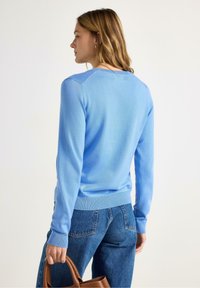 Maglione azzurro chiaro con scollo tondo e bordo a coste. Presenta maniche lunghe e una texture morbida, abbinato a jeans blu scuro e una borsa marrone.