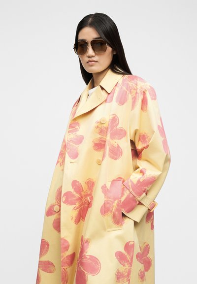 Γυναίκα που φορά φαρδιά κίτρινο παλτό με μεγάλα ροζ λουλουδάτα σχέδια και καφέ γυαλιά ηλίου, ποζάροντας μπροστά σε απλό ανοιχτόχρωμο φόντο.