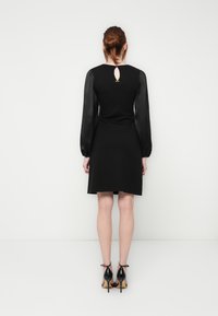 Woman with auburn hair in a low bun wearing a black dress with sheer long sleeves and black high-heeled sandals, standing facing a white wall.