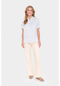 Ljust blå, kortärmad blus med mandarin-krage och knappslå, matchad med krämfärgade byxor med vida ben och mönstrade platta skor.