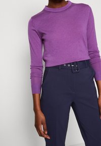 Lila långärmad topp med rund halsringning, tillsammans med marinblå högmidjade byxor med bälte och flera bältesöglor.