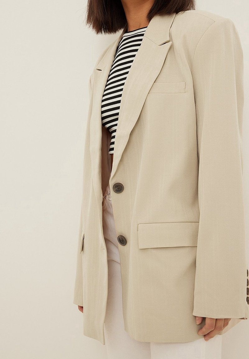 Beige oversized blazer met gestructureerde schouders, twee knopen aan de voorkant en zijzakken. Gedragen over een zwart-wit gestreept shirt.