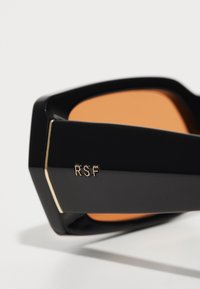 Gafas de sol negras con montura hexagonal, lentes naranjas, detalle del logo RSF en dorado y textura mate suave. El diseño angular realza la estética moderna.