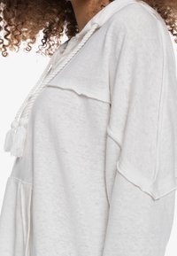 Sudadera blanca con capucha, fabricada en un tejido suave. Presenta un escote en forma de V, cordón decorativo con borlas y mangas anchas.