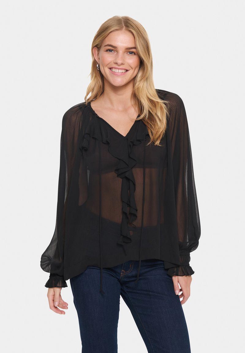 Blusa nera trasparente con dettagli a volant lungo il collo a V e nei polsini. Le maniche lunghe e i dettagli annodati creano una silhouette leggera e fluida.