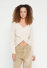 Vrouw met krullend haar, gekleed in een crèmekleurige geribbelde wikkeltrui die de buik blootlaat en beige high-waisted broek, staande tegen een witte achtergrond.