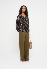 Sort bluse med pink og creme mønster, med V-hals og lange ærmer. Olivengrønne vide bukser. Lysebrun taske, flade sandaler.