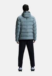 Lichtblauwe pufferjas met een capuchon, met gewatteerde secties, een gladde textuur en verstelbare manchetten, gecombineerd met zwarte broek en schoenen.