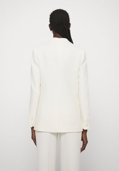 Person med flettet hår iført en hvid skræddersyet blazer og matchende hvide bukser, stående med ryggen til mod en ensfarvet baggrund.