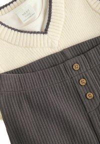 Baby-Outfit mit einem cremefarbenen gerippten Oberteil mit V-Ausschnitt und einem dunkelgrauen gerippten Unterteil. Bemerkenswerte Details sind drei beige Knöpfe am Bund.