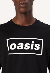 Juodas medvilnės marškinėliai su apvalia apykakle ir trumpomis rankovėmis, kuriuose yra didelis baltas dėžutės logotipas su žodžiu "oasis", užrašytu paryškintu šriftu.