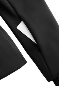 Crna jakna od tkanine s glatkom teksturom i modernim krojem, s bijelom oznakom koja proviruje iz unutarnjeg džepa. Minimalistički dizajn.