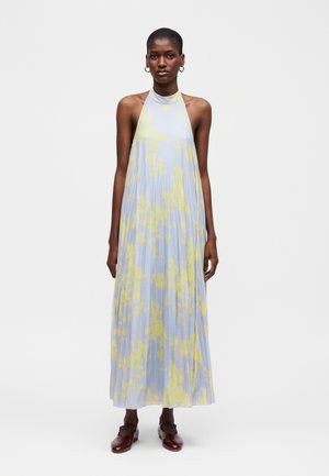 Γυναίκα που φοράει αμάνικο, μακρύ φόρεμα με πτυχώσεις και ψηλό λαιμό, με ανοιχτό μπλε και κίτρινο αφηρημένο λουλουδάτο μοτίβο και σκούρα κόκκινα παπούτσια.