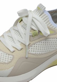Zapatilla ligera en beige y blanco con parte superior de malla, patrones texturizados y cordones planos blancos. Cuenta con una suave suela de espuma y sutiles acentos.