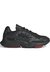 Scarpa sportiva nera in materiale mesh e sintetico, con tre strisce prominenti, superficie testurizzata e suola rossa.