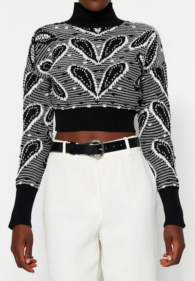 Pull à col roulé noir et blanc avec des motifs abstraits de feuilles, associé à un pantalon blanc taille haute et une ceinture noire à boucle argentée.