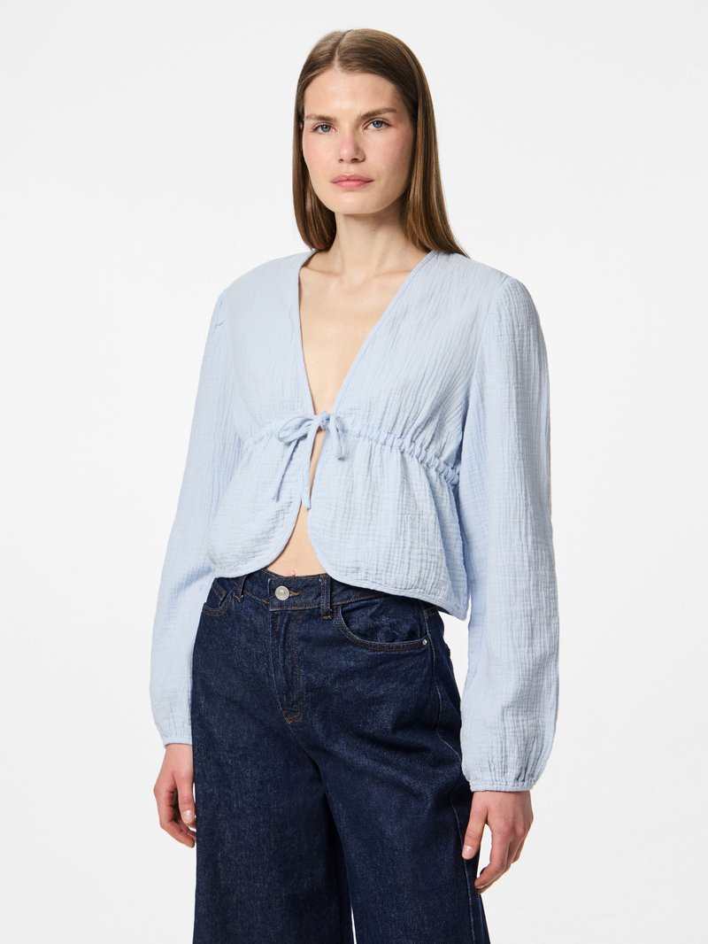 Femme aux cheveux bruns et lisses portant un haut bleu clair à manches longues, col en V avec nœud devant et un jean bleu foncé taille haute, debout calmement.