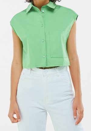 Camisa recortada sin mangas de color verde claro con botones y bolsillo en el pecho, combinada con vaqueros blancos de tiro alto, en una persona de pie.