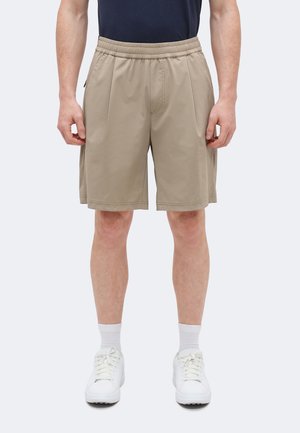 Muškarac obučen u bež kratke hlače s elastičnim strukom, bijele čarape i bijele tenisice, stoji nasuprot jednobojnoj svijetloj pozadini.