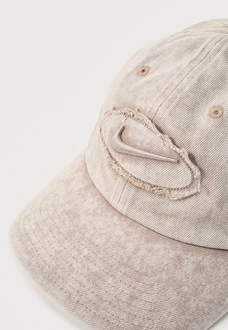 Gorra de béisbol beige con un parche ovalado desgastado y un logo swoosh curvado cosido en el panel frontal, mostrada sobre un fondo blanco.
