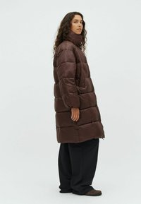 Cappotto puffo marrone con colletto alto, design trapuntato e vestibilità rilassata. Indossato sopra pantaloni neri larghi, dotati di tasche laterali e superficie liscia.