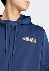 Námornícky modré zip-up mikina vyrobená z hladkej látky, s výrazným logom Adidas so žltým okrajom na ľavej hrudi a nastaviteľnou kapucňou.