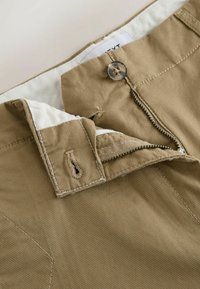 Pantalon beige avec une fermeture éclair partiellement ouverte, un bouton fixé au-dessus et une doublure intérieure blanche visible.