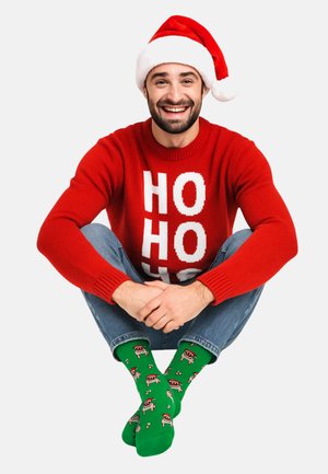 Red sweater with white "HO HO HO" text, blue jeans, and green socks featuring patterned holiday designs, paired with a white and red Santa hat.