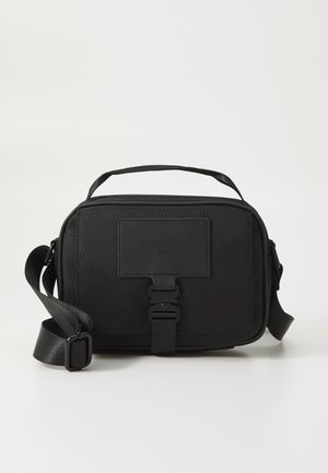 Petit sac bandoulière noir compact avec sangle réglable, poignée supérieure, fermeture à boucle avant et patch rectangulaire sur une surface en tissu texturé.