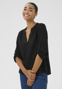 Blouse noire à boutons avec un col en V, manches retroussées et tissu texturé, associée à un jean bleu, présentée sur un fond neutre.
