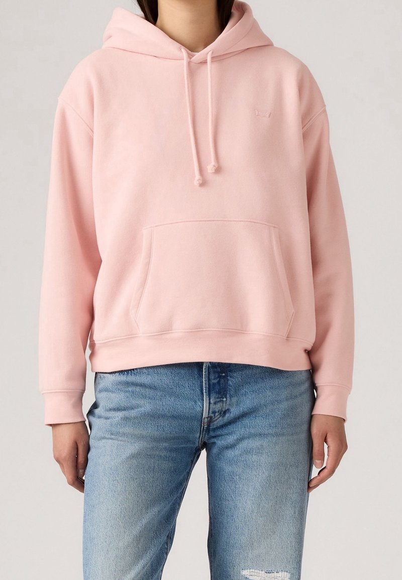 Personne portant un sweat à capuche rose clair avec poche avant et un jean bleu légèrement usé.
