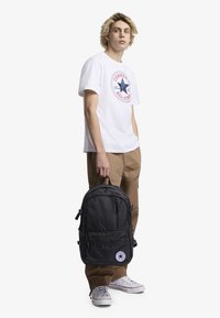 Mochila de tela negra con múltiples compartimentos con cremallera, que cuenta con un parche de logo circular. El modelo lleva una camiseta gráfica blanca y pantalones beige.