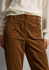 Braune Corduroyhosen mit einer strukturierten Oberfläche, ausgestattet mit zwei Gesäßtaschen und einer hüfthohen Taille, kombiniert mit einem hellcremigen Pullover.
