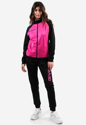Donna che indossa una giacca a zip Givova rosa e nera e pantaloni neri con il logo Givova rosa, in piedi su un fondo neutro con scarpe da ginnastica bianche.