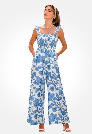 Frau trägt einen weißen Overall mit blauem Blumenmuster, Rüschen-Trägern, Perlenkette und weißen Sandalen mit hohen Absätzen, steht mit den Händen in den Taschen.