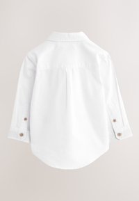 Vue de dos d'une chemise blanche à manches longues avec poignets boutonnés et un pli central dans le dos sur un fond uni.