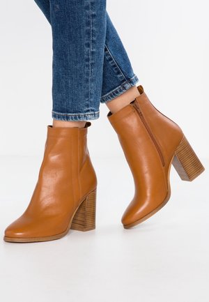 Bottes en cuir marron à la cheville avec un bout arrondi, une fermeture éclair latérale et un talon épais à motif. Portées avec un jean bleu raccourci.